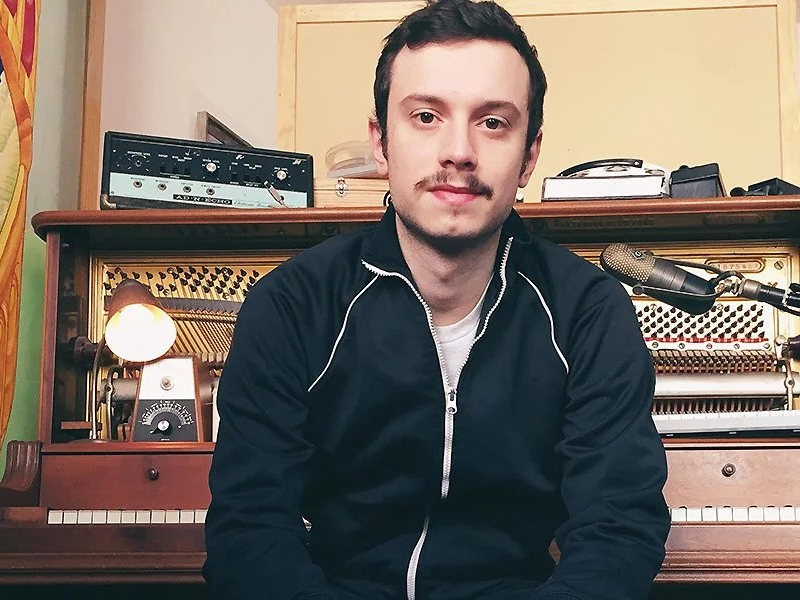 Joe sits in front of a piano and microphone. He wears a black jacket with white zip and a moustache