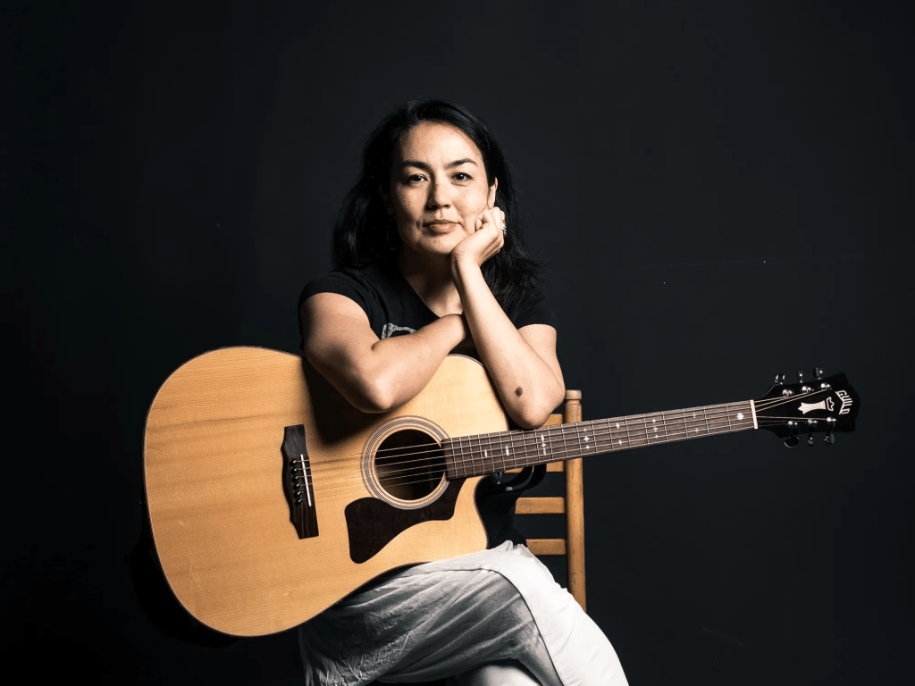 Jasmine sits leaning on her acoustic guitar which rests in her lap. She is half smiling. Her left eyebrow is slightly raised