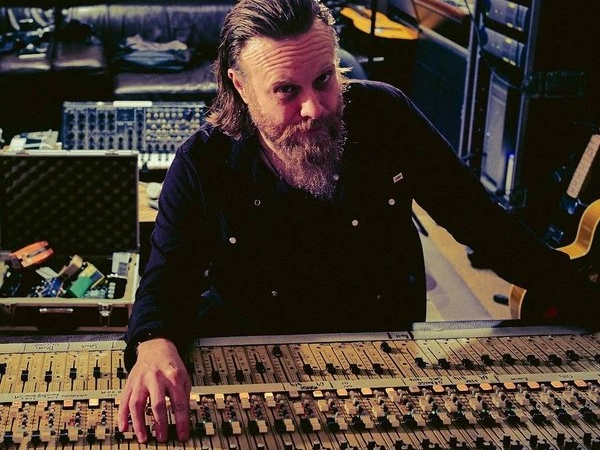Dave leans over a mixing desk looking to camera. He has a full beard and moustache
