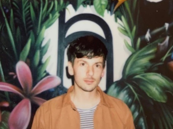 Martin has dark hair and stubble. He looks to camera wearing a stripy t shirt under a beige shirt